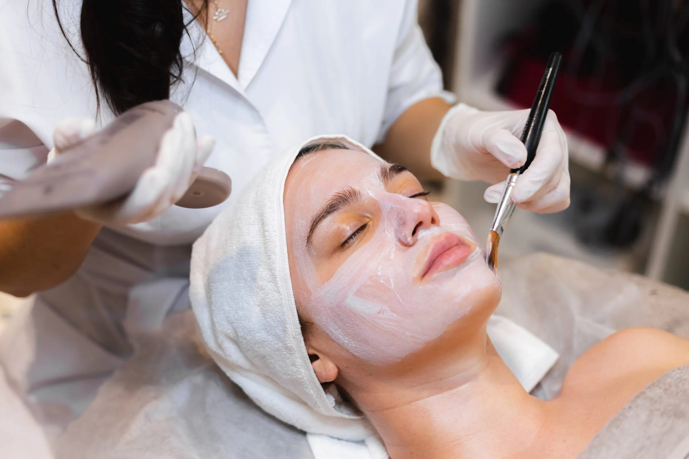 Gloved esthetician brushing a mask onto a woman wearing a towel turban